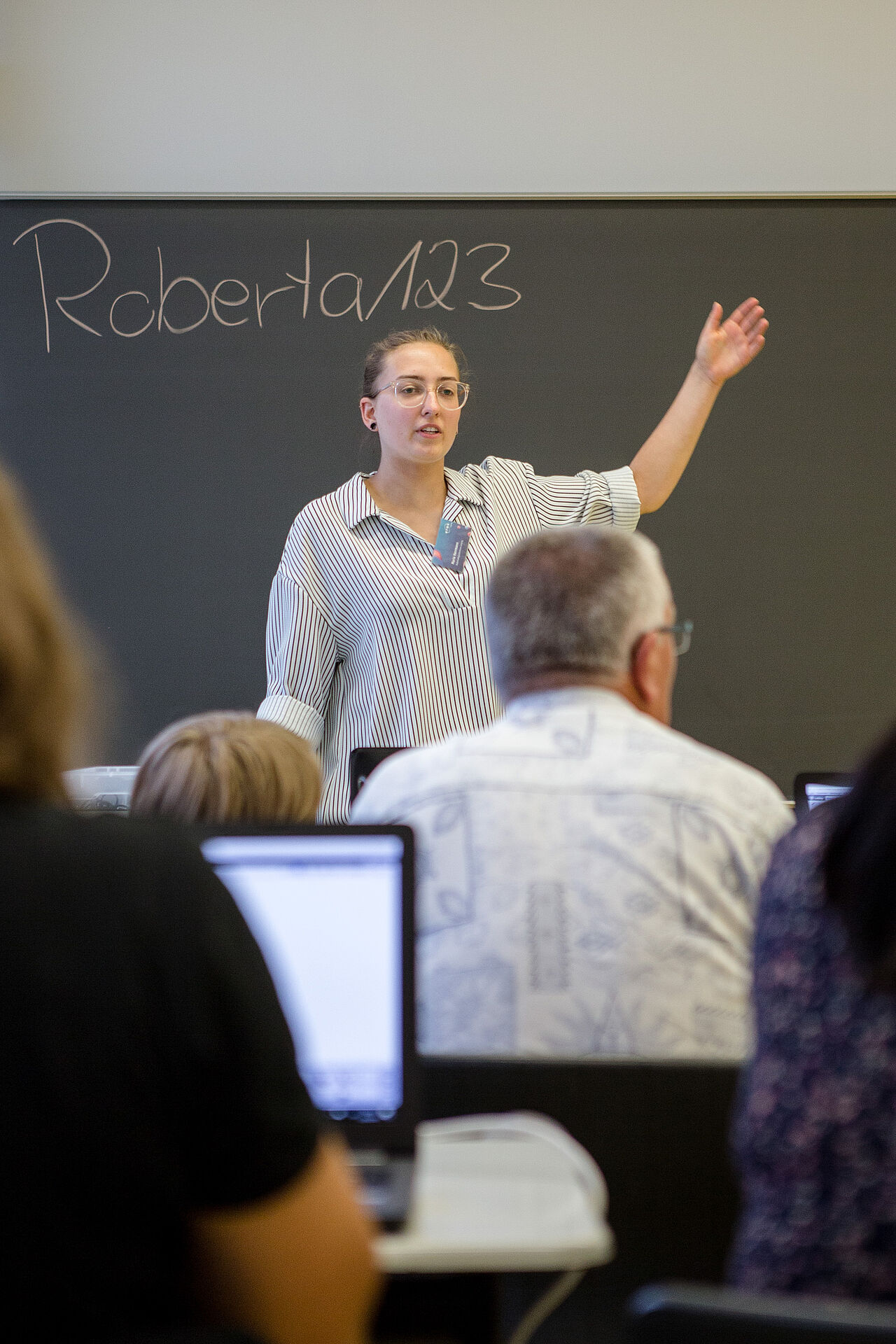 Eine junge Frau steht vorne im Seminarraum und zeigt auf die Tafel, auf der das Passwort zum Starten der Computer steht.
